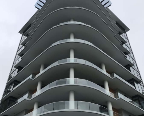 A modern multi-story building with curved balconies and glass railings, viewed from below. The top floor features distinctive vertical slats extending upward, and the structure has a sleek, contemporary design.