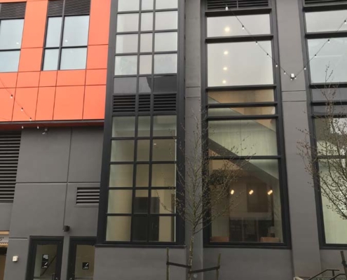 Modern building facade with large vertical windows, orange and gray wall panels, two small doors at the base, bare young trees, string lights above, and steps leading up to an entrance on the right.