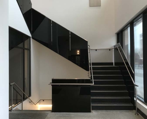 A modern indoor staircase with black railings and steps, surrounded by large windows letting in natural light, and polished concrete floors. The design is minimalistic and features clean lines and neutral colors.