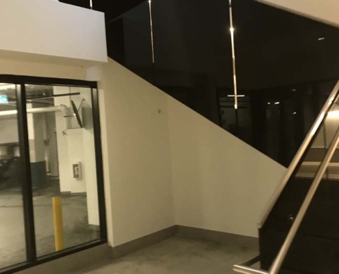 A modern indoor stairwell with black railings and glossy black paneling, next to a large window revealing a parking garage; walls and floors are light-colored, giving a minimalist look.
