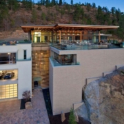 Modern multi-level house built into a hillside, featuring large glass windows, a rooftop pool, balconies, and a garage with sports cars visible, surrounded by trees and rocky landscape at dusk.