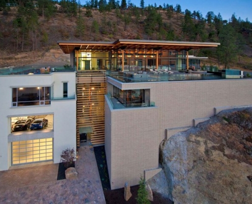 Modern multi-level house built into a hillside, featuring large glass windows, a rooftop pool, balconies, and a garage with sports cars visible, surrounded by trees and rocky landscape at dusk.