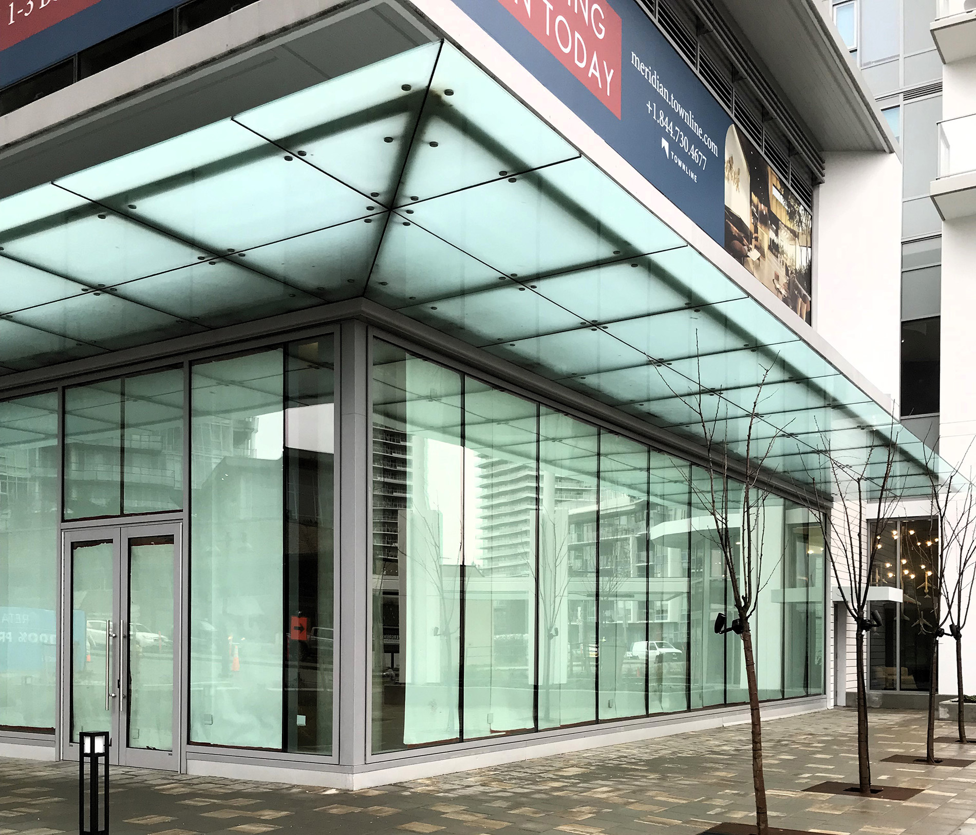 A modern glass storefront on a city street corner with large floor-to-ceiling windows, double glass doors, and a glass canopy above. Leafless trees and buildings are visible outside.
