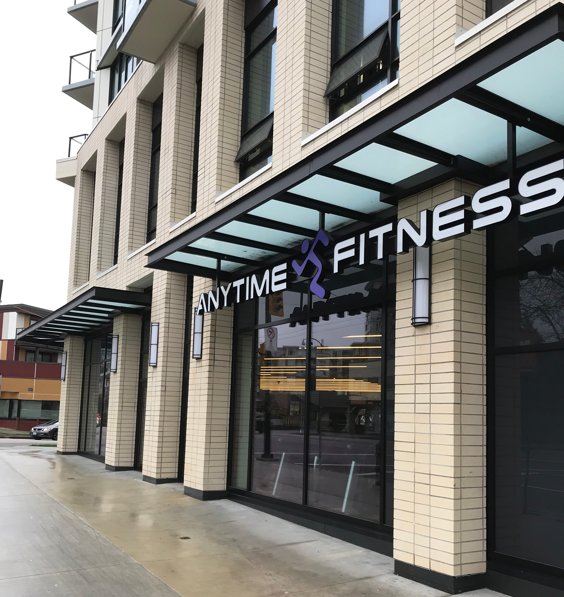 Street view of an Anytime Fitness gym with cream brick exterior, large glass windows, and a glass awning. The Anytime Fitness sign with a running figure logo is visible above the entrance.