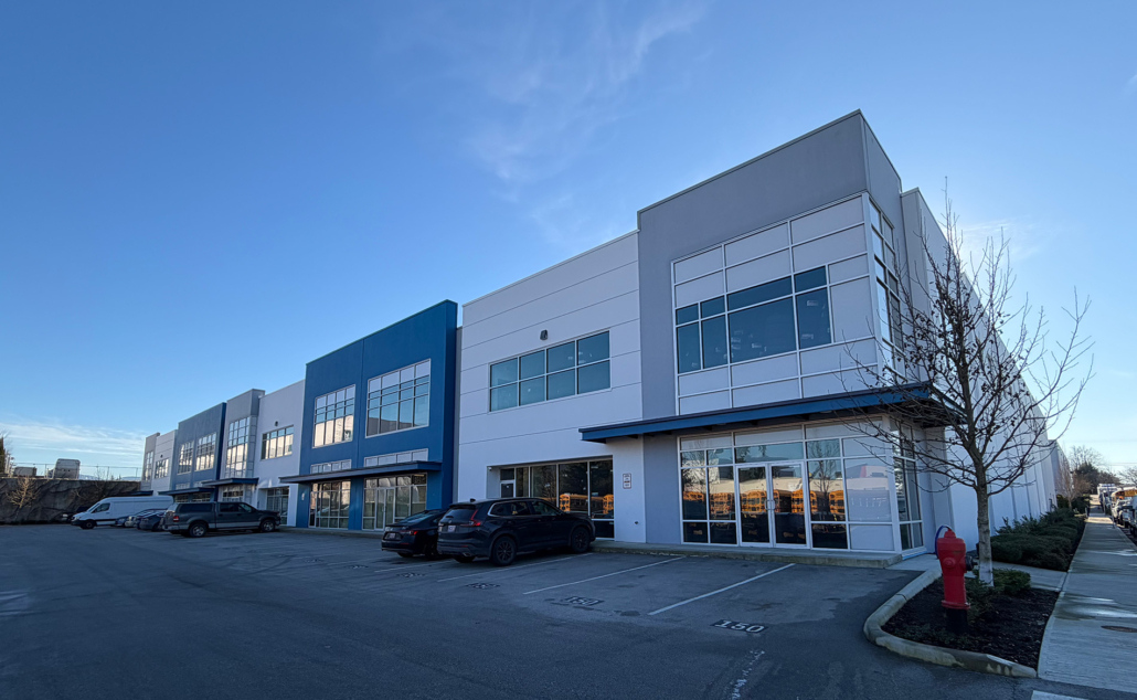 A modern commercial building with large windows, multiple storefronts, and parked cars in front. The sky is clear and blue, and a leafless tree and red fire hydrant are visible near the entrance.