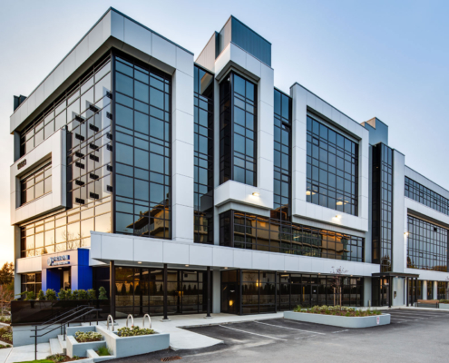 A modern three-story office building with large glass windows, white trim, and black accents. The parking lot in front is mostly empty, and the sky is clear with the sun setting in the background.