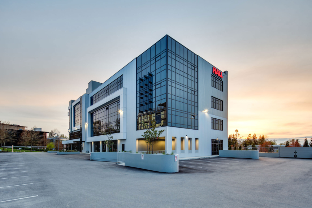 A modern, three-story office building with large glass windows and the red HAO logo on the top corner, set against a sunset sky with an empty parking lot in front.