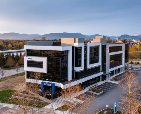 A modern, glass-fronted office building with geometric architecture is surrounded by trees and parking lots, with mountains visible in the background under a clear sky.
