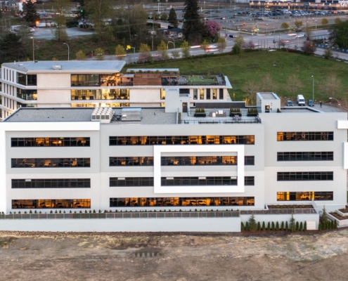 A modern four-story office building with large windows, surrounded by parking lots, green spaces, and trees, with city streets and more buildings visible in the background.