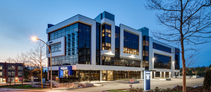 A modern, multi-story office building with large glass windows and geometric design, illuminated at dusk. The parking lot in front is mostly empty, and a few trees border the property.