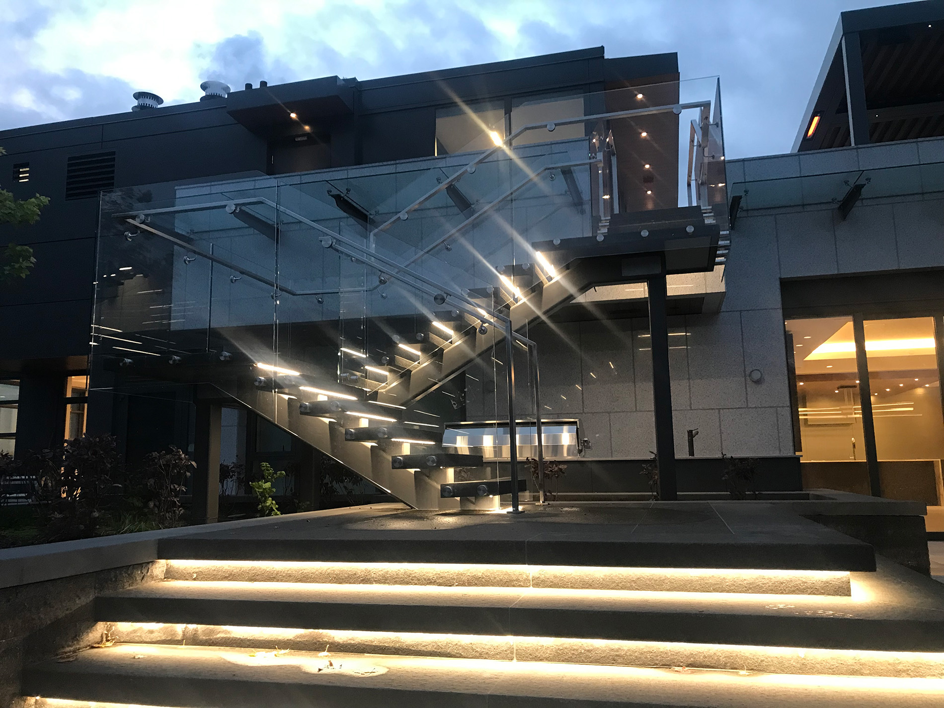 A modern outdoor staircase with glass railings and built-in lights leads to the upper level of a contemporary building, illuminated in the evening.
