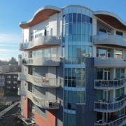 A modern apartment building with curved balconies and large glass windows, set against a clear blue sky. Other residential buildings and trees are visible in the background.