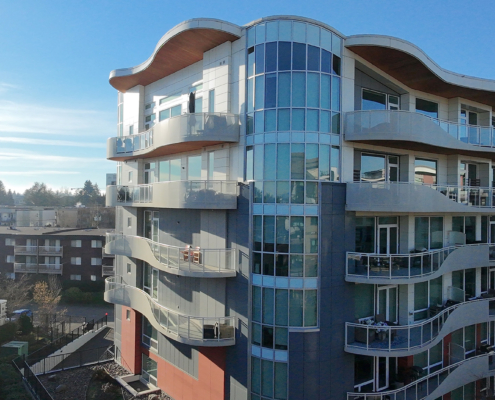 A modern apartment building with curved balconies and large glass windows, set against a clear blue sky. Other residential buildings and trees are visible in the background.