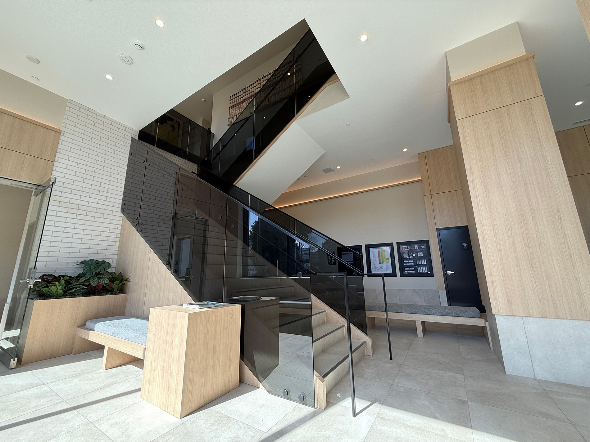 A modern lobby features light wood benches, cream-colored tiles, and a central black glass staircase. Large windows and indoor plants add brightness and freshness to the stylish, minimalist space.