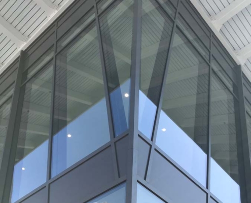 A modern glass building is seen from below, reflecting a tall skyscraper. The image shows sharp architectural lines, large windows, and a white ceiling with beams above.