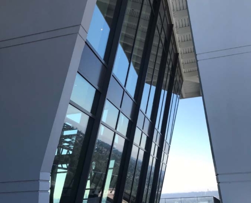 Tall, modern glass windows slant outward on a white building, reflecting the sky. Below, a row of tables and chairs sits on a terrace with a view of a distant landscape.