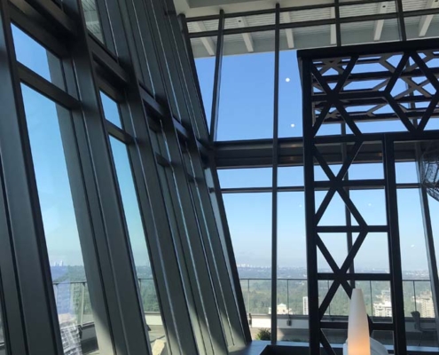 Large floor-to-ceiling windows with angled glass panels overlook a distant landscape under a blue sky; inside, there is a modern black metal structure and a tall, slender, glowing lamp.