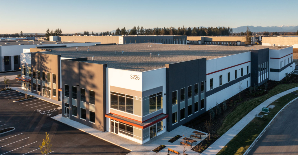 Modern industrial warehouse building with large windows, geometric design, and the number 3225 displayed. Surrounded by parking spaces, sidewalks, landscaped areas, and other industrial buildings under clear skies.