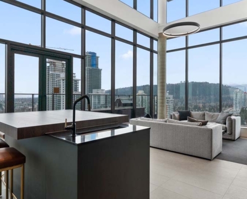Modern high-rise apartment with floor-to-ceiling windows, stylish kitchen island with a black countertop, gray sofas, and views of city buildings and forested hills in the background.
