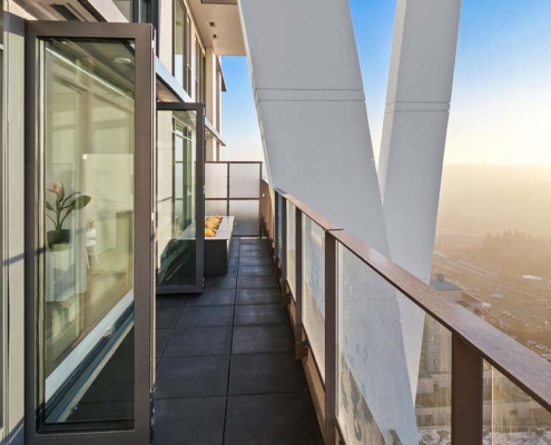Modern high-rise balcony with glass railing, large structural supports, and open glass door. Cityscape is visible through mist in the background on a sunny day.