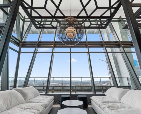 Modern lounge area with two white sofas facing each other, geometric black-framed ceiling, large windows offering a cityscape view, and a unique pendant light hanging in the center.