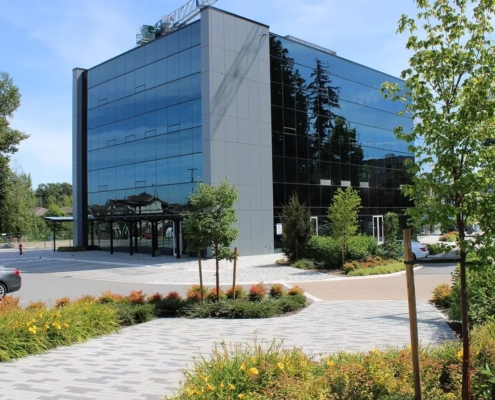 A modern, glass-fronted office building surrounded by landscaped gardens, young trees, and paved walkways on a sunny day. A parked car is visible on the left.