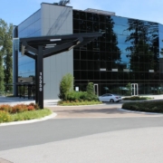 A modern glass office building with reflective windows is surrounded by trees and greenery. A silver car is parked nearby, and a black street sign displays the number 15350 at the entrance.