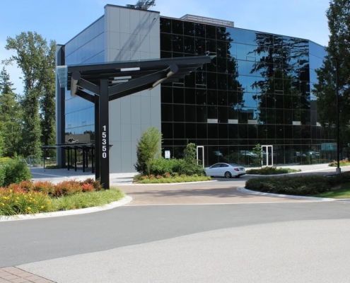 A modern glass office building with reflective windows is surrounded by trees and greenery. A silver car is parked nearby, and a black street sign displays the number 15350 at the entrance.