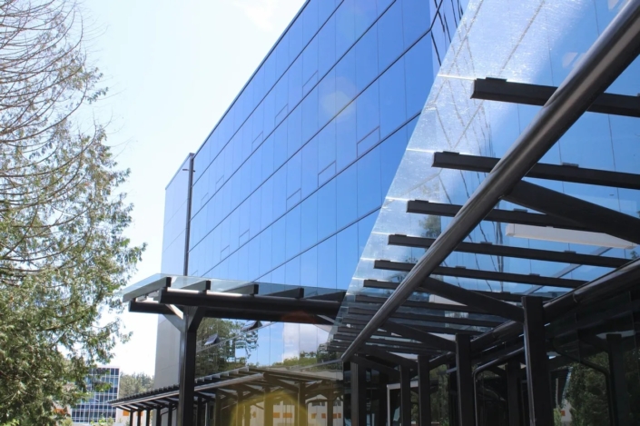 A modern glass office building with reflective blue windows and a black metal and glass awning, surrounded by trees and a clear sky.