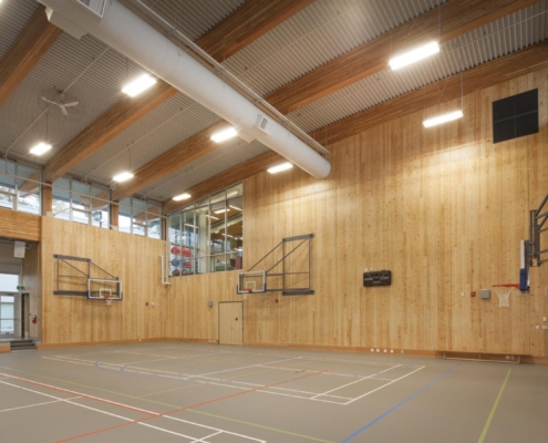 A modern indoor gymnasium with tall wooden walls, high windows, basketball hoops, and a gray floor marked with colorful sports court lines. The space is well-lit with overhead lights.