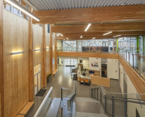 A spacious modern building interior with large wooden beams, glass railings, high ceilings, and natural light from many windows. There are desks, chairs, and a reception area on the ground floor.