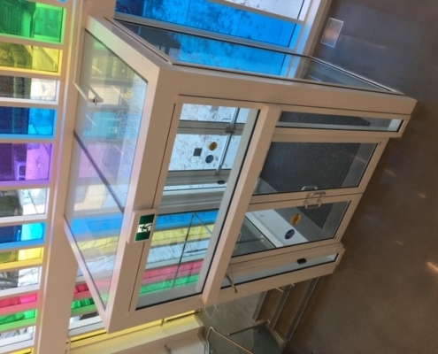 A modern glass entrance with double doors and colorful, tinted window panels above and to the side, casting rainbow reflections inside the building. Snow is visible outside through the doors.