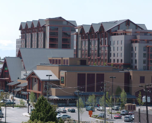 A large multi-story building complex with steeply pitched roofs, red trim, and multiple sections, surrounded by trees and a parking lot under a clear sky.