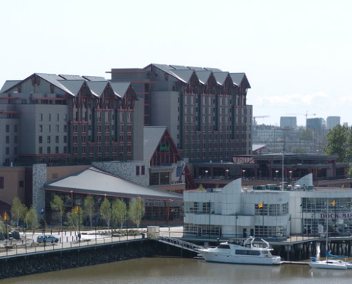 A large waterfront hotel with multiple peaked roofs sits behind a marina with docked boats and a white building labeled Dock Marina, surrounded by trees and a river.