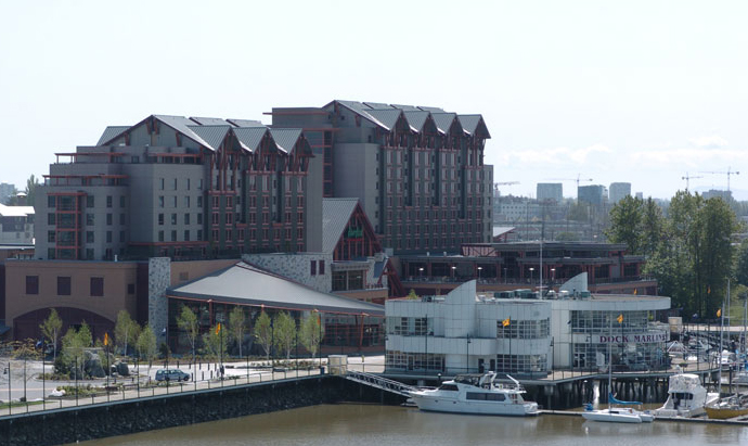 A large waterfront hotel with multiple peaked roofs sits behind a marina with docked boats and a white building labeled Dock Marina, surrounded by trees and a river.