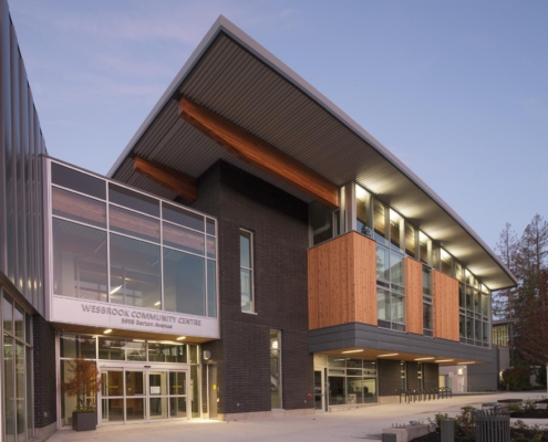 Modern building with large windows, angled roof, and wooden accents. The entrance reads Wesbrook Community Centre above glass doors, and the exterior is a mix of dark brick and light wood panels. Trees are visible in the background.