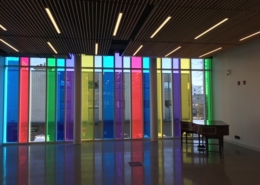 A modern interior with floor-to-ceiling windows featuring vertical panels of red, blue, yellow, green, and purple. Sunlight shines through the colorful glass, casting vibrant reflections on the shiny floor. A wooden desk sits by the wall.