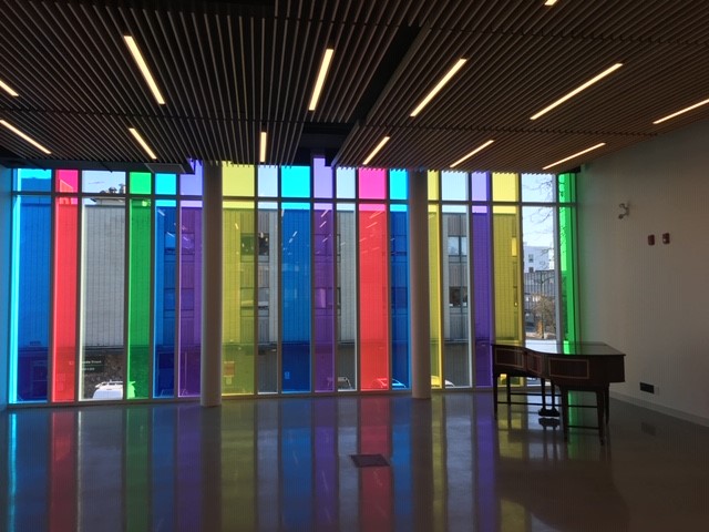 A modern interior with floor-to-ceiling windows featuring vertical panels of red, blue, yellow, green, and purple. Sunlight shines through the colorful glass, casting vibrant reflections on the shiny floor. A wooden desk sits by the wall.