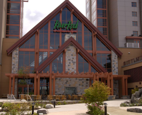 The image shows the front entrance of the River Rock Casino Resort, a modern building with large glass windows, stone accents, and tall red-brown beams, surrounded by landscaped greenery and outdoor seating.
