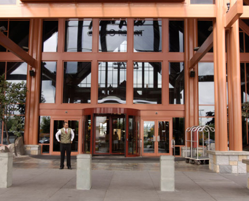 A formally dressed attendant stands outside the large glass entrance of a modern building with tall orange beams and stone accents. A luggage cart is nearby, and the area is clean and spacious.
