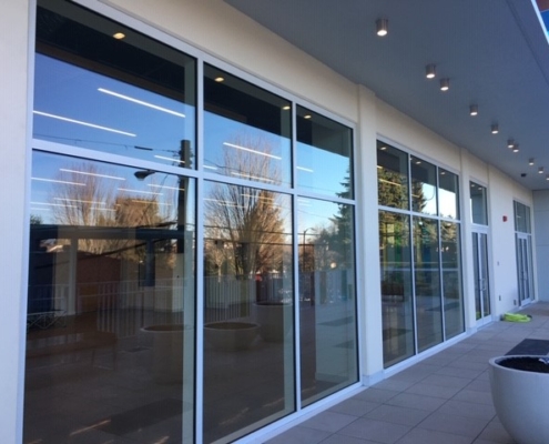 Large glass windows reflect trees, sky, and nearby buildings along a modern buildings exterior. The patio area is tiled, with ceiling lights and a large planter in the foreground.
