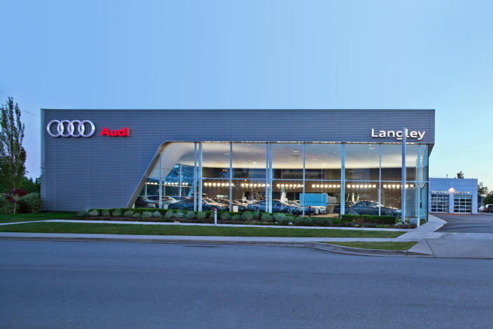 Modern Audi dealership building with large glass windows and prominent Audi and Langley signage. Several cars are visible inside the well-lit showroom, and a service center is attached on the right side of the building.