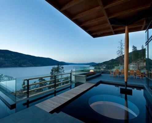 Modern outdoor terrace with glass railings, an infinity pool, circular hot tub, wooden deck, and seating area overlooking a calm lake and tree-covered hills at sunset.