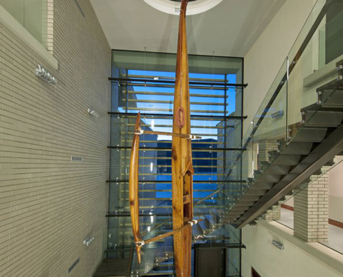 Large wooden canoe sculptures hang in the open atrium of a modern building with glass walls, metal staircases, and a circular skylight above, allowing blue evening light to spill in.
