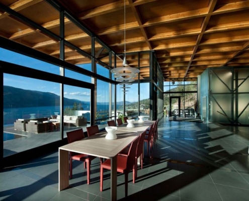 Modern dining area with floor-to-ceiling glass windows, wooden ceiling, red chairs, and a large table. The room overlooks a scenic lake and mountains, with outdoor seating visible on the patio.