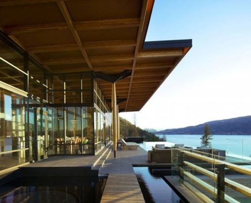A modern building with large glass walls overlooks a lake and mountains. A wooden walkway and an outdoor pool run alongside the building, with lounge chairs arranged near a glass railing. The sky is clear and blue.
