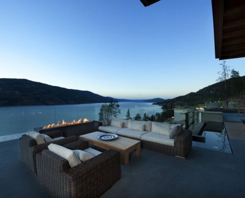 Modern outdoor patio with wicker furniture and fire pit overlooks a serene lake and mountains at dusk, with clear sky and calm water reflecting the landscape.