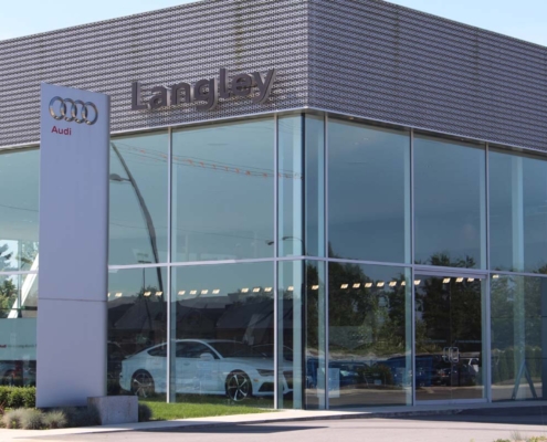 Modern glass-walled Audi dealership building with Langley signage, showcasing cars inside. An Audi sign stands outside, and the sky and trees are reflected in the windows.