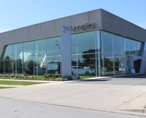 Modern Audi car dealership with large glass windows, displaying several vehicles inside. The exterior features a sleek, metallic design with the Audi logo and signage visible. A paved driveway and sidewalk are in front.