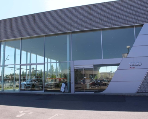 Exterior view of a modern Audi car dealership with large glass windows, showing cars displayed inside. The building has a sleek design with an Audi logo and sign visible on the right side.